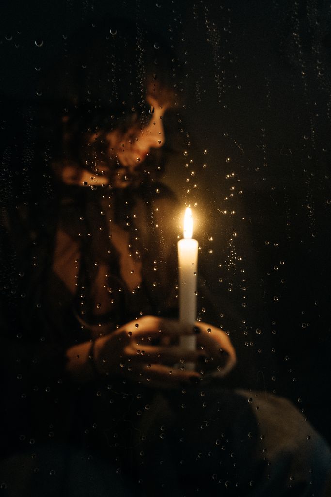 Moody portrait of a person with a candle seen through rain-covered glass.